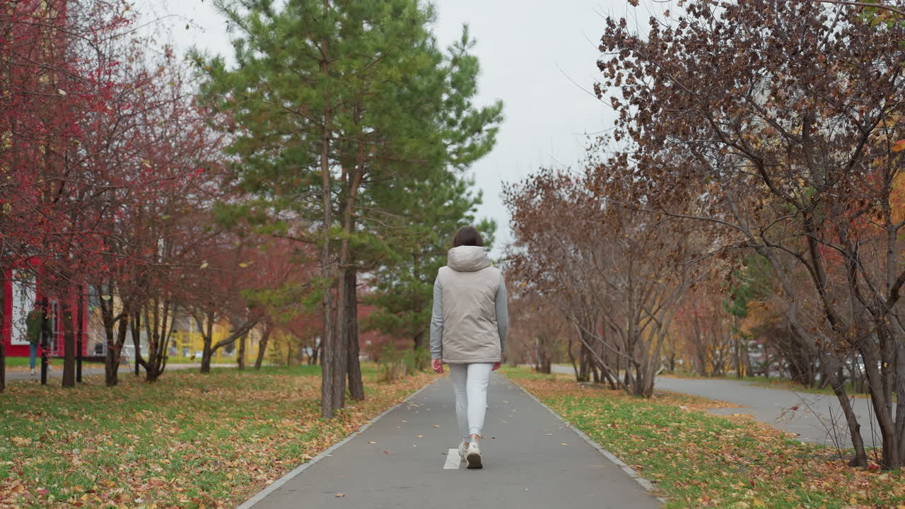 Runner dressed in light jacket and joggers walks along marked path surrounded by autumn trees swaying in gentle breeze while car moves in distance on serene day in peaceful urban park setting