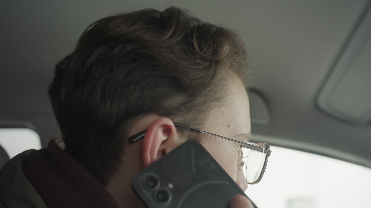 close up of young boy wearing glasses changing phone to left hand while on call inside car, prepares to start vehicle by inserting key, cold winter light filtering through window