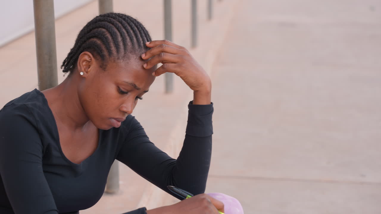 young woman sitting outdoors studying stressed, holding head over notebook, pen poised, anxious expression, braided hair, urban sidewalk setting, casual clothing, tense posture, exam preparation mood