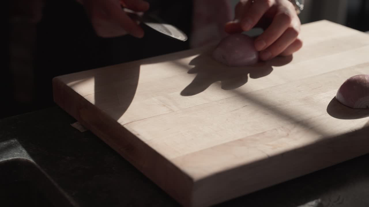 En japansk kokkekniv løftes fra et solbeskinnet skærebræt for at tilberede et skalotteløg (slowmotion 4K)