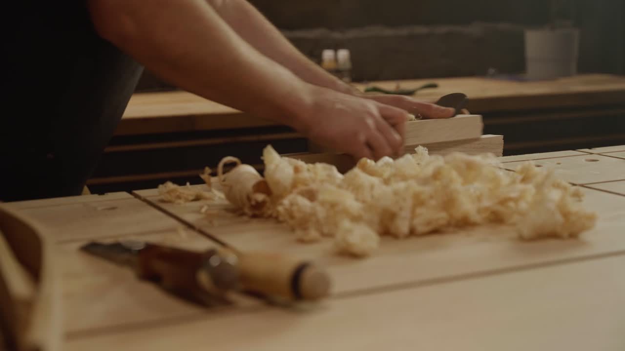 Close-up of a craftsman’s hands planing a wooden board with wood shavings scattered on the workbench, symbolizing craftsmanship, precision, and traditional woodworking