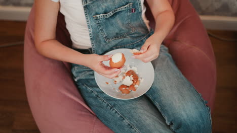 top down view of person wearing blue denim overall sitting on pink bean bag, carefully peeling brown egg into white bowl filled with broken shells, relaxed indoor setting on wooden floor