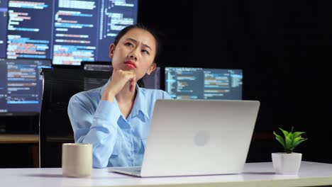 programadora asiática pensando en algo y levantando el dedo índice mientras escribe código en una computadora portátil usando múltiples monitores que muestran la base de datos en los escritorios de la oficina