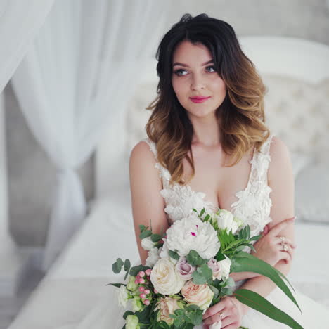 attractive young bride in a sexy wedding dress is sitting on bed. beautiful bride with flowers. pretty model in white dress is posing to the female photographer on the bedroom background.