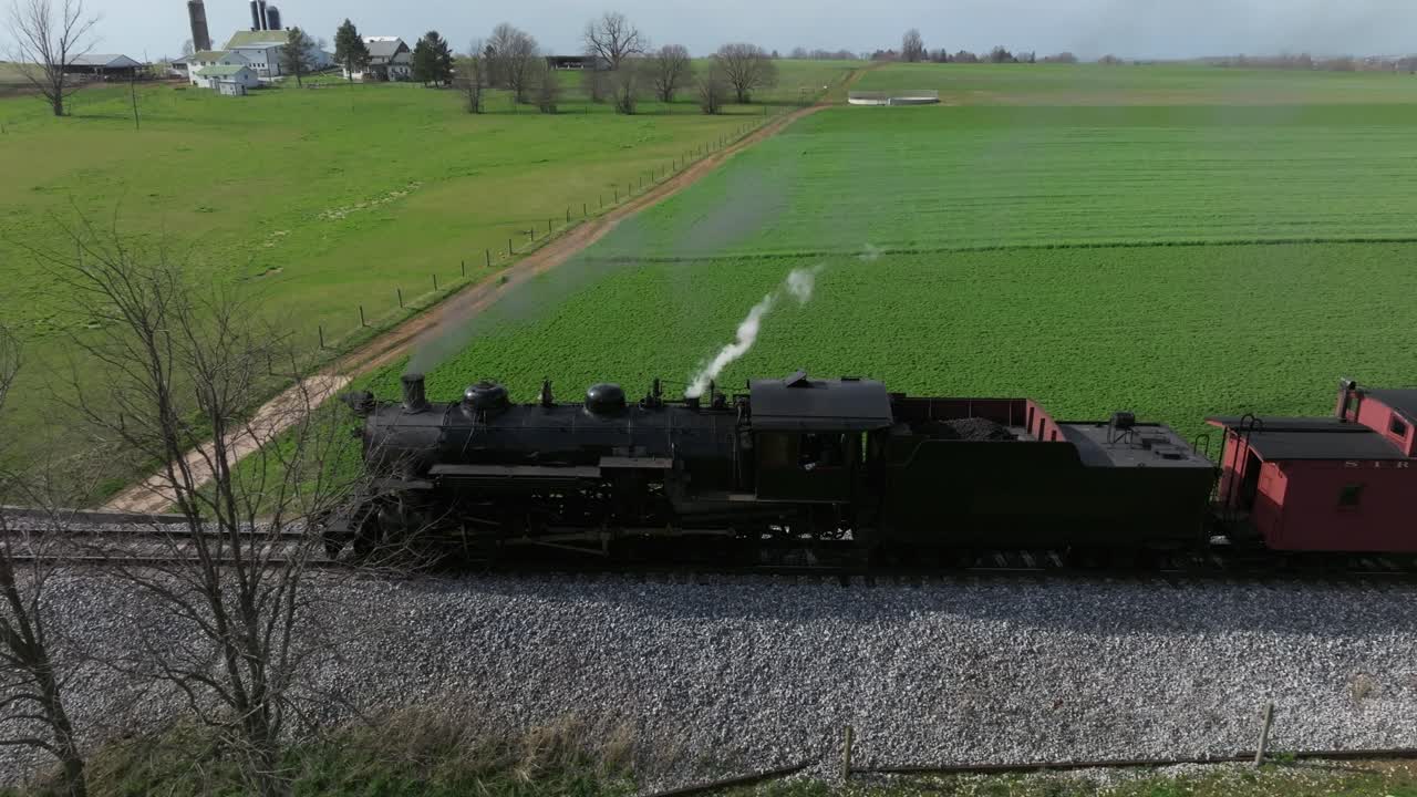 A vintage steam train chugs along the tracks, surrounded by vibrant green fields and trees under a clear sky. Its whistle pierces the calm, evoking nostalgia.
