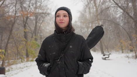 lady in black hoodie carrying guitar in pack with hands in pockets, standing thoughtfully in snowfall on a snow-covered path, surrounded by bare trees and serene winter scenery