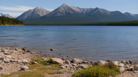 a serene landscape featuring a vast lake with a rocky shore, surrounded by dense forests and majestic snow-capped mountains under a clear blue sky