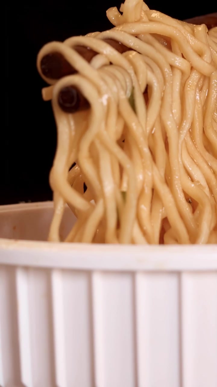Le bacchette afferrano e sollevano noodles istantanei cotti da una tazza bianca su uno sfondo scuro, con dettagli in primo piano e illuminazione da studio uniforme.