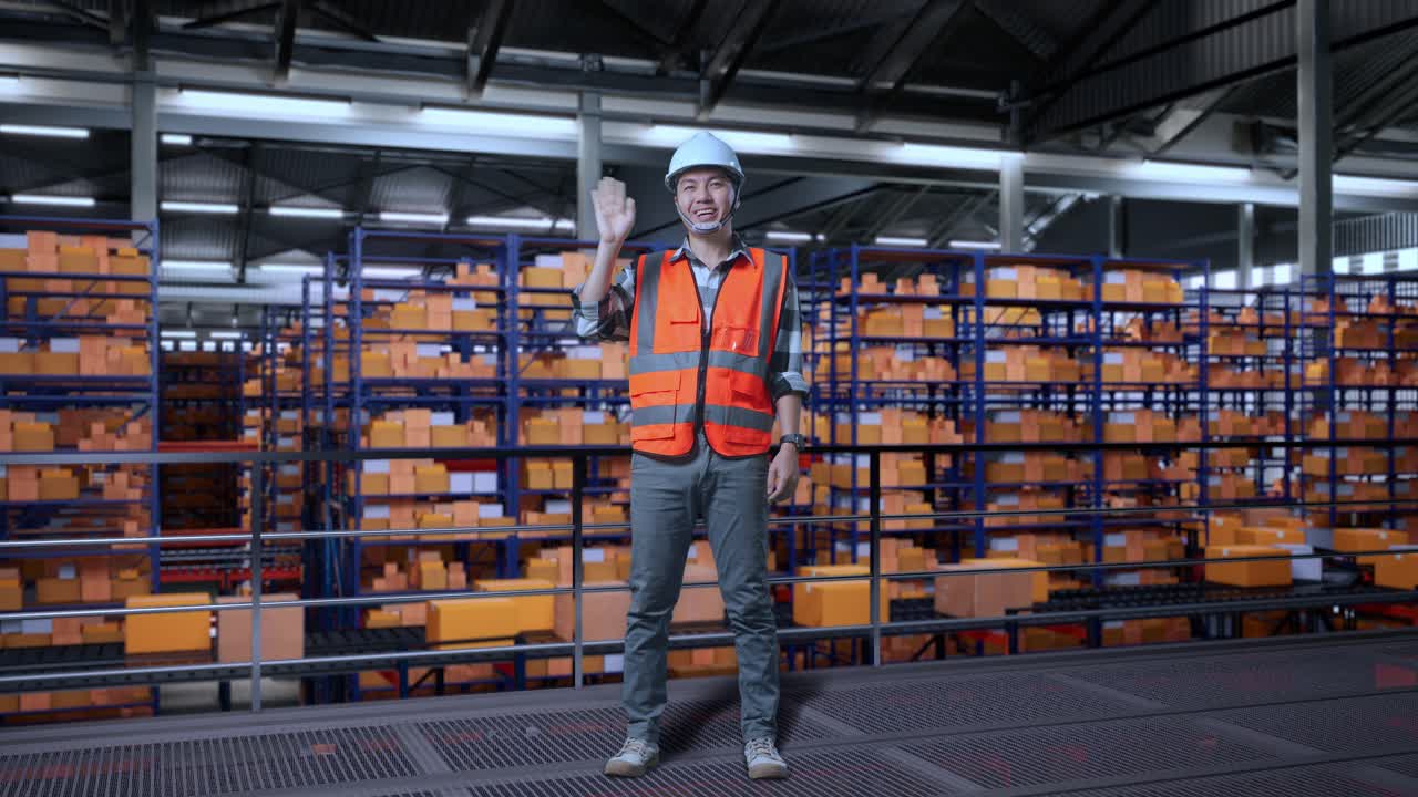 Full Body Of Asian Male Engineer With Safety Helmet Standing In The Warehouse With Shelves Full Of Delivery Goods. Smiling To Camera And Waving Hand Saying Good Bye In The Storage