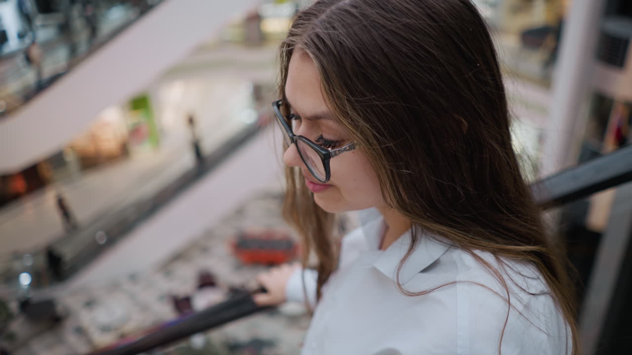 primer plano de una mujer con gafas en una escalera mecánica en un centro comercial concurrido, en un estado de ánimo reflexivo mientras mira hacia la derecha, capturando un momento de contemplación en medio del ajetreo
