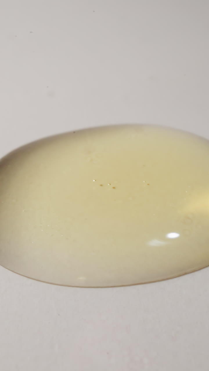 Heavy drops of fresh jojoba oil fall on table reflecting light. Transparent and shiny liquid spreads on plain white surface macro