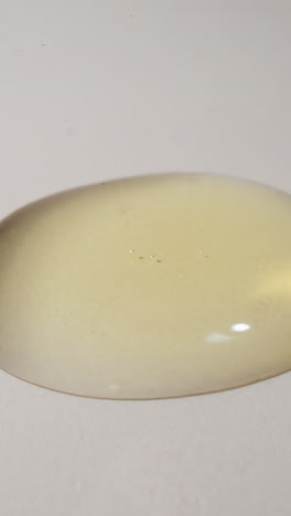 Heavy drops of fresh jojoba oil fall on table reflecting light. Transparent and shiny liquid spreads on plain white surface macro
