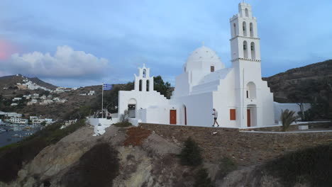 Drone-footage-of-man-walking-next-to-white-church-on-the-island-Ios,-Greece-near-Ios-port-in-Chora