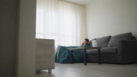 boy covered in blanket coughs while lying on sofa in bright modern room, covering mouth with hand, resting quietly beside tissue box on table, suggesting cold symptoms