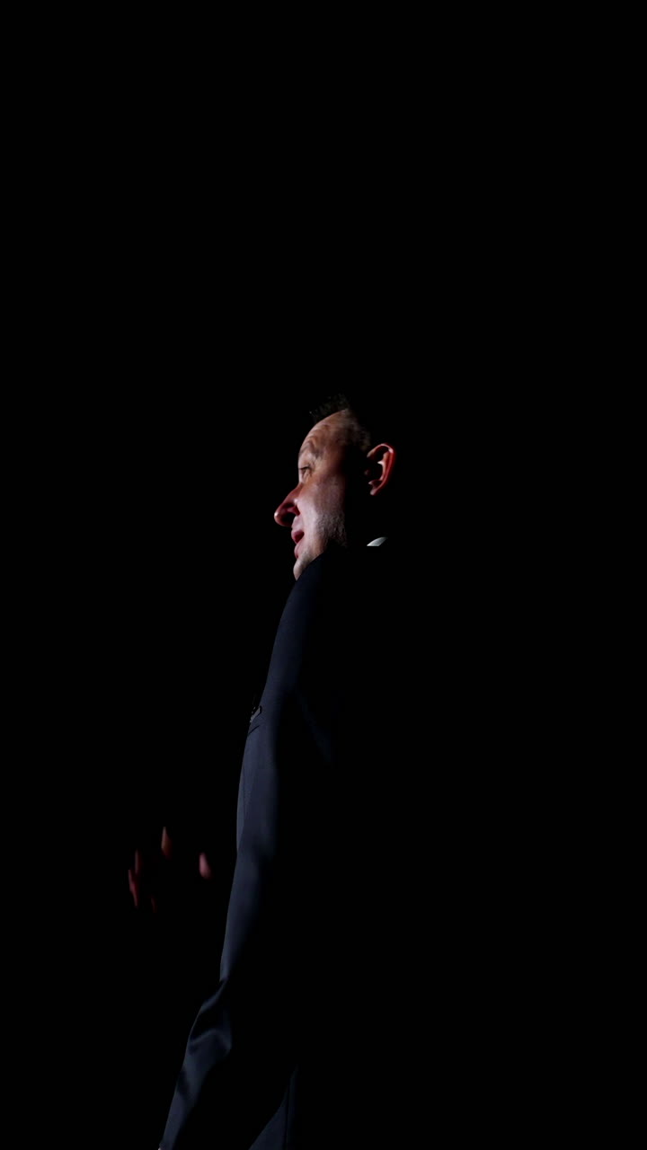 Speaker on black background. Profile view of a man talking to the audience while standing on stage. Performer in darkness. Vertical video