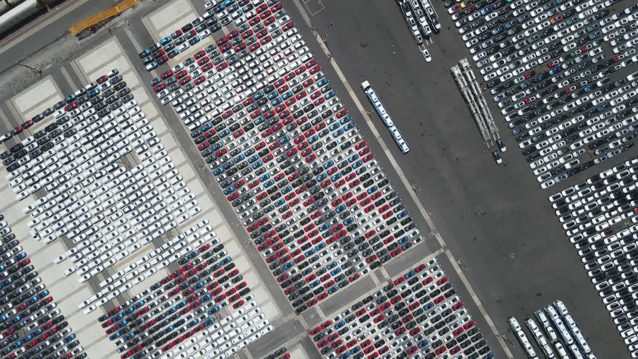 A sweeping drone view features a massive lot filled with tightly packed cars in a range of colors, showcasing the scale and symmetry of vehicle storage.