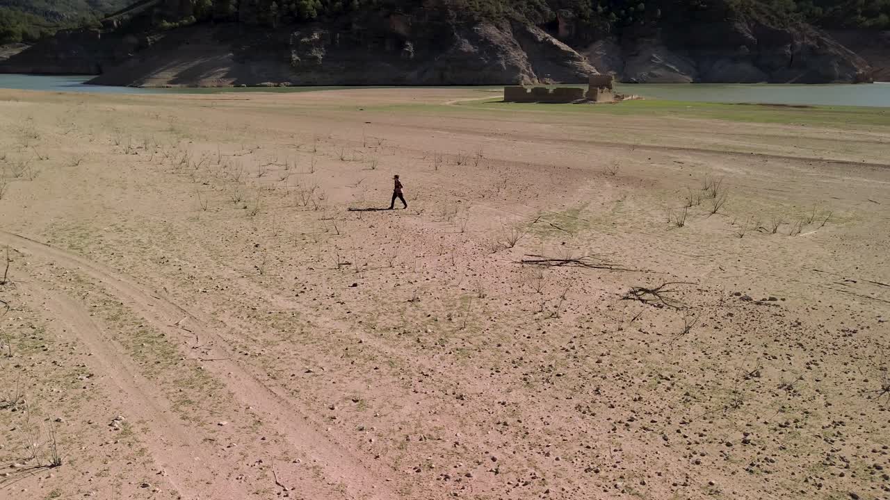 Drone footage of a solitary woman wandering along the shores of a marsh, near ruins, in a parched and desolate landscape
