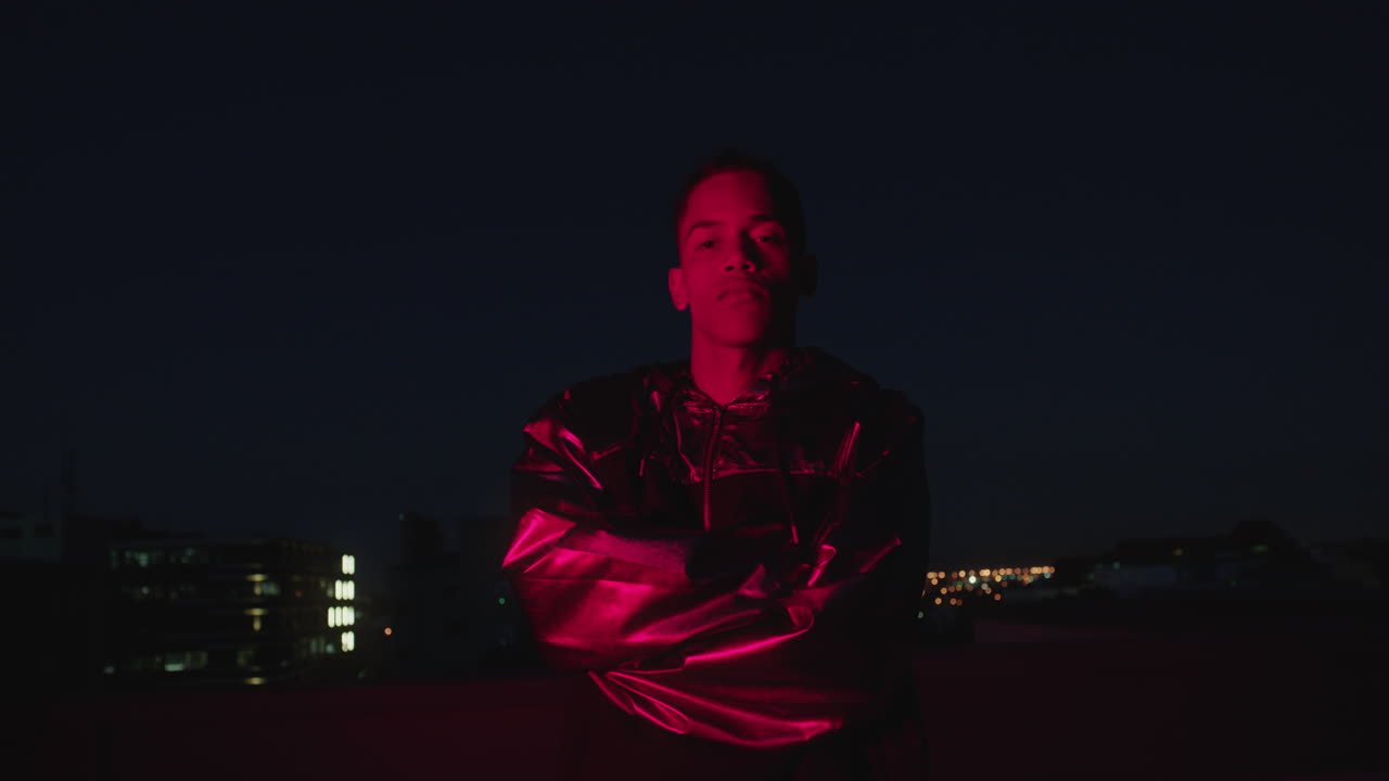 портрет человека смешанной расы с перекрещенными руками, позирующего в городе ночью, уверенно глядя на красный свет