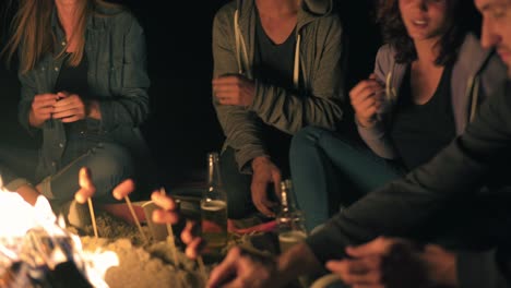 Group-of-young-friends-sitting-by-the-fire-late-at-night,-grilling-sausages-and-drinking-beer,-talking-and-having-fun