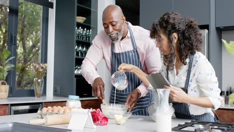 Happy-biracial-couple-wearing-aprons-baking-and-using-tablet-in-kitchen,-slow-motion