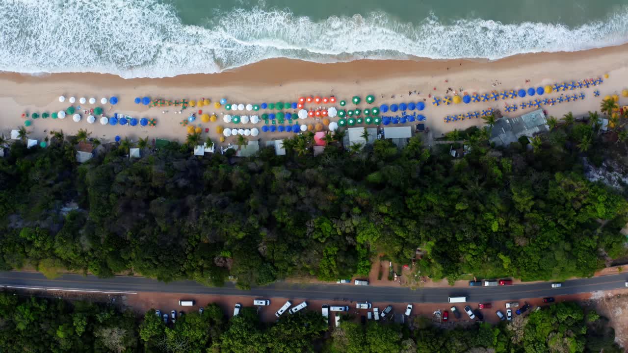 vista aérea de drones a vista de pájaro de una pequeña carretera rodeada de árboles tópicos y autos estacionados con la playa de praia do madeiro cerca de pipa con turistas, sombrillas y arena dorada en el norte de brasil