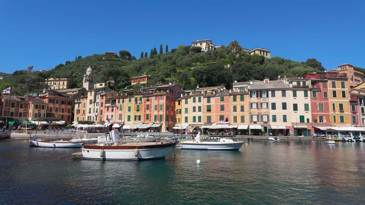 Ampia inquadratura vivace di Portofino, l'iconico porto d'Italia con colorati edifici sul lungomare, barche ormeggiate, una rigogliosa collina verde e acqua scintillante sotto una luce diurna chiara e brillante