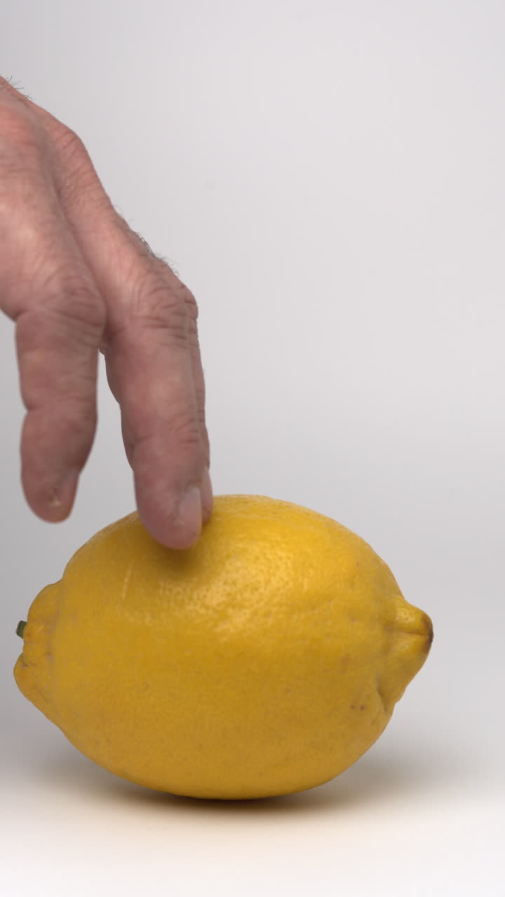 Video al rallentatore della mano di un uomo che muove delicatamente un limone da un lato all'altro nella parte inferiore dell'inquadratura, come se stesse pensando a cosa farne, su uno sfondo bianco pulito.
