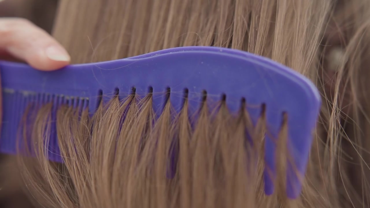 Vue latérale rapprochée d'un peigne violet à dents larges passant à travers de longs cheveux châtain clair, avec une séparation visible des mèches, un léger volume et une action de démêlage en douceur, une main tenant le peigne sur un fond sombre