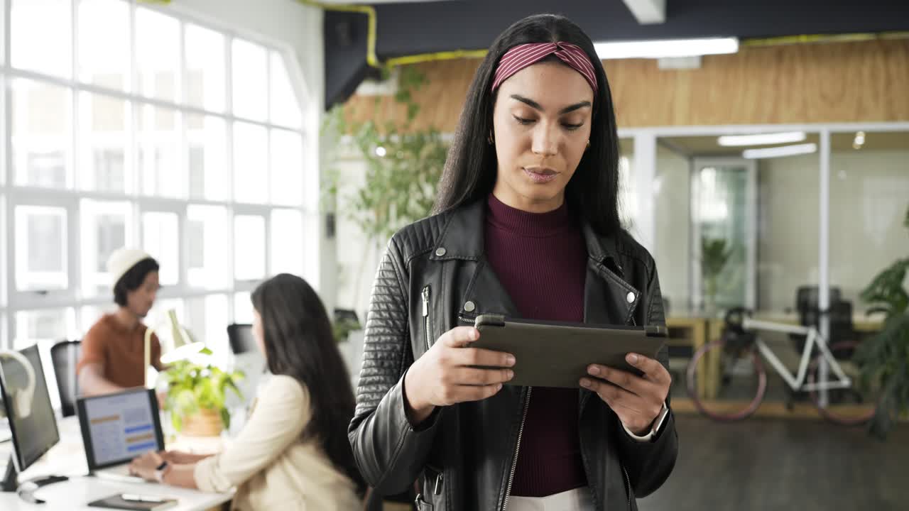 Female office worker in open-plan workspace tapping tablet and reviewing report before smiling