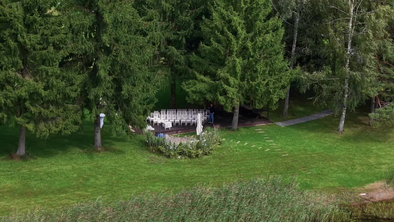 Aerial footage captures a secluded outdoor wedding ceremony setup in a forest clearing by a lakeshore, with white chairs, lush greenery, and serene natural scenery bathed in daylight.