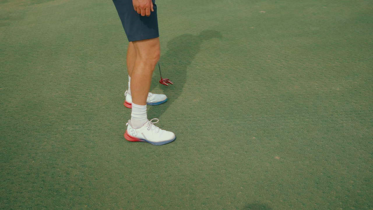 Slow-motion clip showing a male golfer from the waist down walking across the putting green with a red putter, to read the green. Capturing calm preparation, rhythm, and detail ahead of a putt.