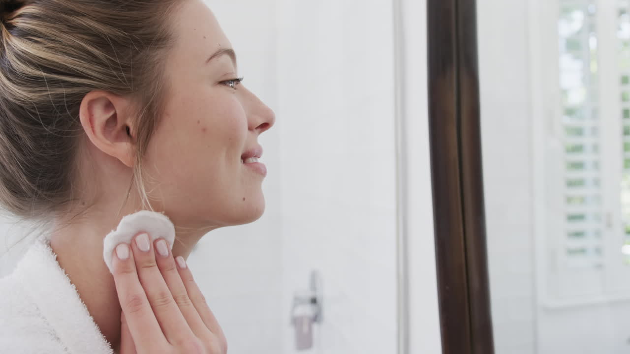 Happy biracial woman cleansing face using cotton pad looking in mirror in bathroom, slow motion
