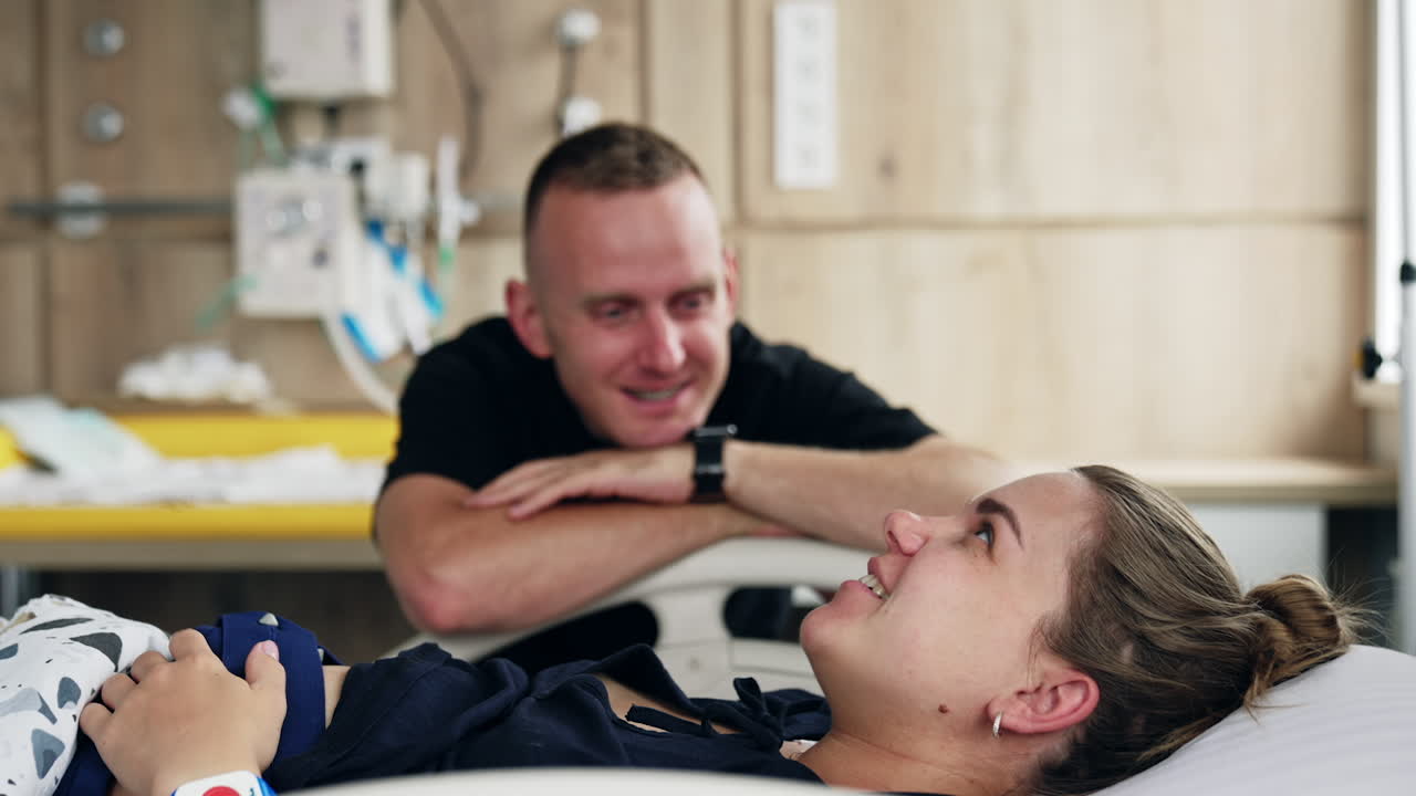 Happy expecting couple in the hospital ward. Man sits next to his wife lying in bed. Expecting spouses talking and laughing.