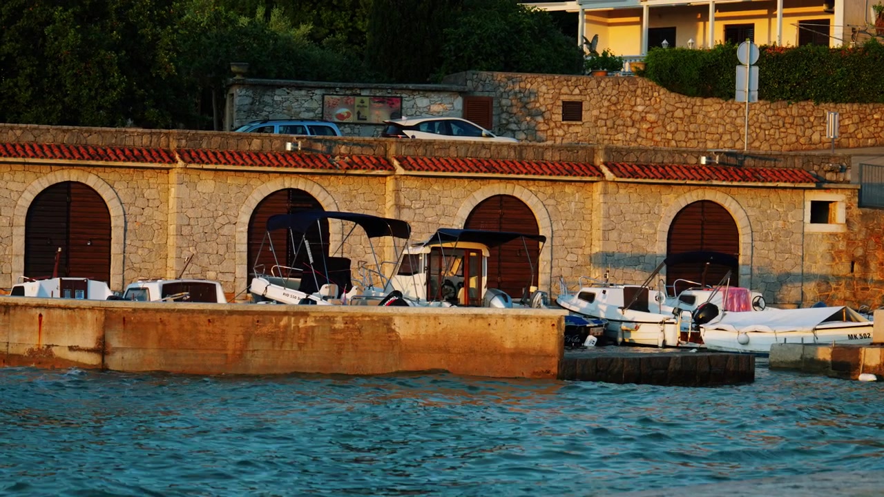 Piccole barche tradizionali ormeggiate accanto all'architettura in pietra del lungomare sull'isola di Krk in Croazia durante la calda luce del tramonto lungo la costa adriatica
