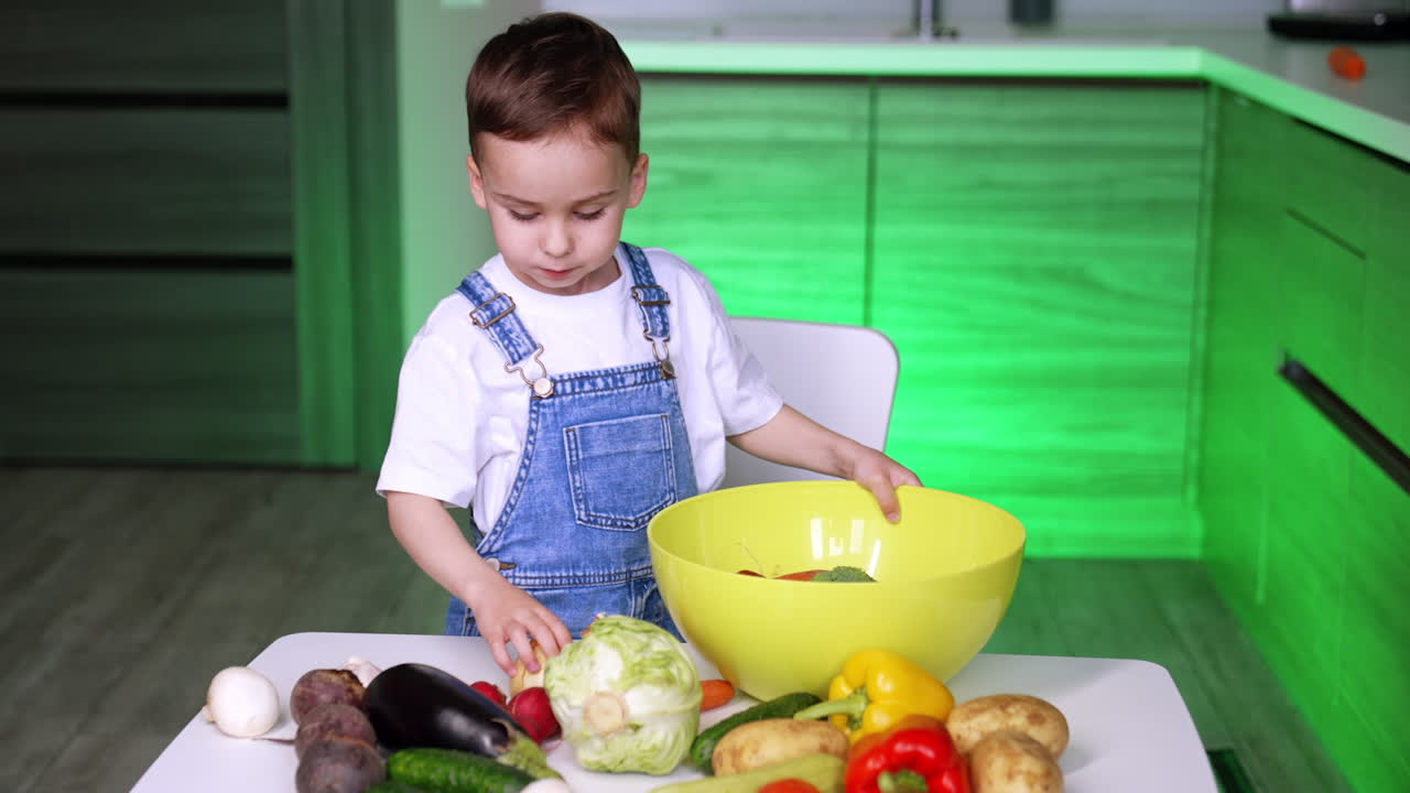 Bambino prepara verdure per cucinare. Un bambino in salopette blu mette verdure fresche in una grande ciotola gialla su un tavolo da cucina.