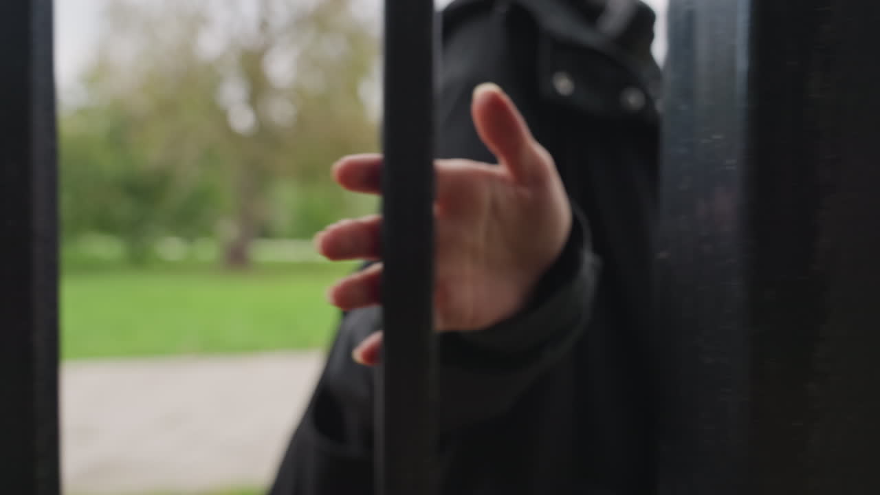 Mano che si allunga attraverso un cancello di ferro, dita che sfiorano sbarre di metallo nero, persona anonima in un cappotto scuro, sentiero del parco e prato verde sfocati sullo sfondo, luce nuvolosa, primo piano intimo che trasmette