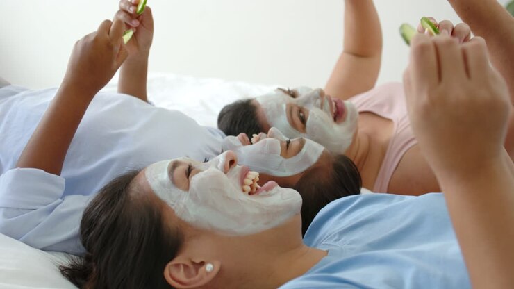 Diverse female friends laughing playfully, lifting cucumbers on bed with facial masks for spa night