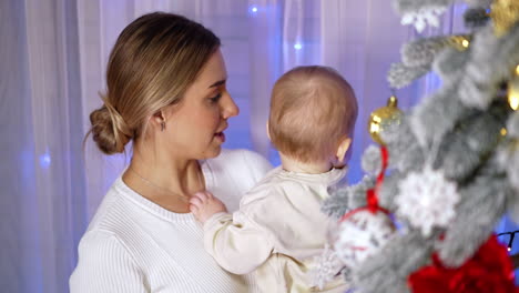 blonde woman in white dress holds a baby standing at the christmas tree. mommy shows the decorations to her son.