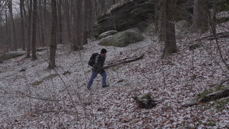 movimiento lento de un excursionista con barba y con una camisa de franela camina por una colina boscosa a principios del invierno con árboles estériles y un polvo de nieve en el suelo