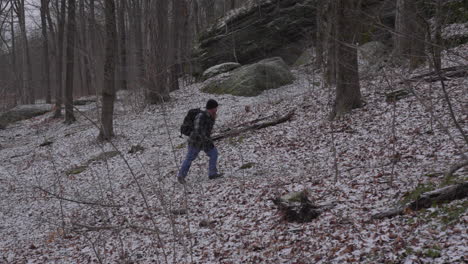 slow motion of a hiker with a beard and wearing a flannel shirt walks up a woodland hill in early winter with barren trees and a dusting of snow on the ground