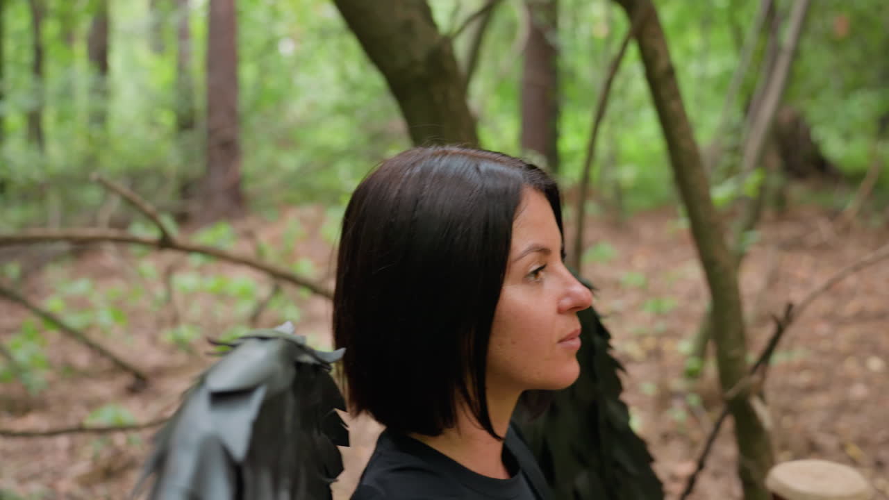 Side view of mysterious woman with black wings walking confidently through green forest, surrounded by soft light and branches, expressing strength, calmness, and enigmatic elegance