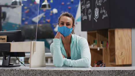 Kaukasische-Frau-Mit-Gesichtsmaske,-Die-An-Der-Rezeption-Der-Indoor-Kletterhalle-Auf-Dem-Tresen-Lehnt