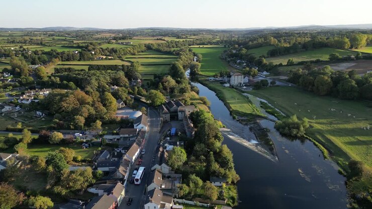 Aerial drone view of Bennettsbridge village along River Nore showing stone mill, flowing water, surrounding farmland, and green countryside under soft daylight sky