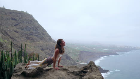 la mujer sentada en el borde de un acantilado en la pose del perro con vistas al océano, respirar el aire del mar durante un viaje de yoga a través de las islas