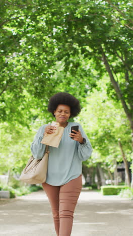 vertical video of african american woman walking through park, using smartphone and eating sandwich