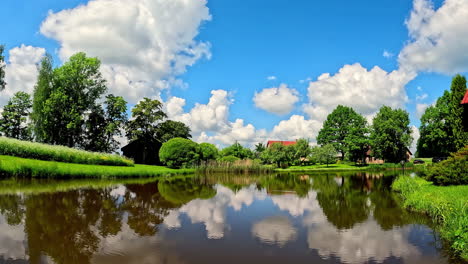 Grandes-Nubes-Esponjosas-Ondeando-Sobre-Una-Cabaña-Y-Reflejándose-En-La-Superficie-De-Un-Estanque---Lapso-De-Tiempo