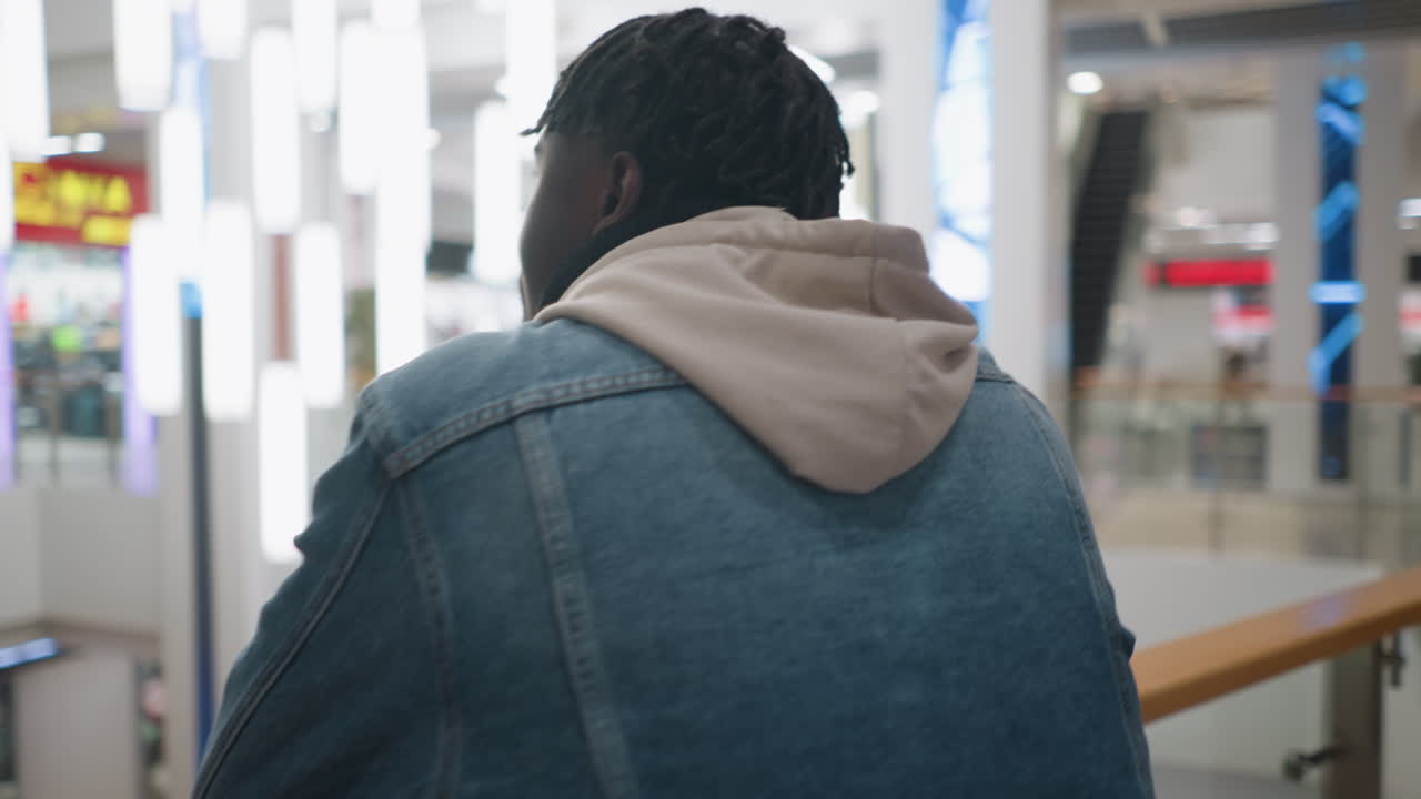Young man in denim jacket and hoodie sipping takeaway coffee while leaning on railing in modern shopping mall, surrounded by glass, store signs, bright ceiling lights, and soft ambient reflections in background