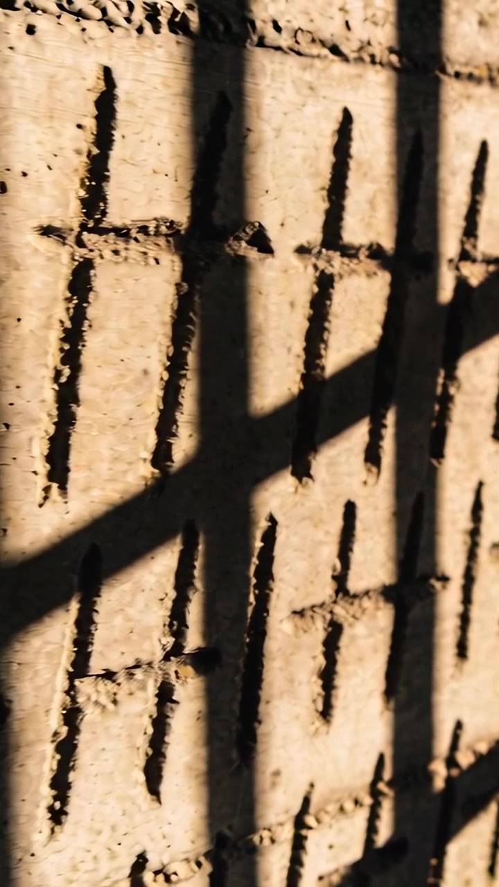 Vertical video: Sun through bars casting slant shadow on wall with vertical slots, camera panning
