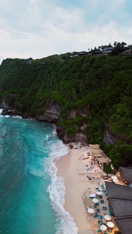 this vertical drone shot showcases a stunning beachfront restaurant in uluwatu, where white sand meets turquoise waters and a massive cliff provides a breathtaking natural backdrop.
