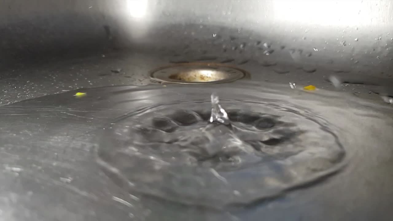 Faucet drainage hole in stainless kitchen sink with water dripping and splashing droplets in slow motion